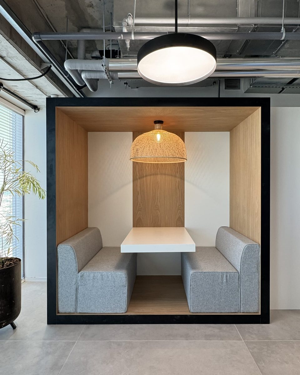 A finished office breakout booth: timber-lined with a black frame, upholstered benches and a rattan pendant over the table